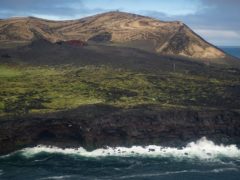 a volcanic island with a grassy meadow among lava plains, with ocean waves crashing against cliffs