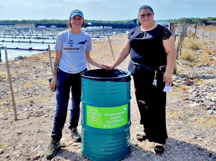Two women stand in the sun of a salt farm with a metal barrel.