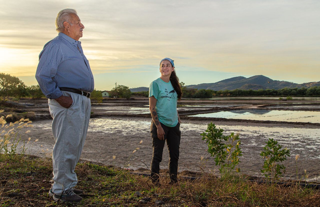 Older man and a woman stand on the shore of a muddy wetland.