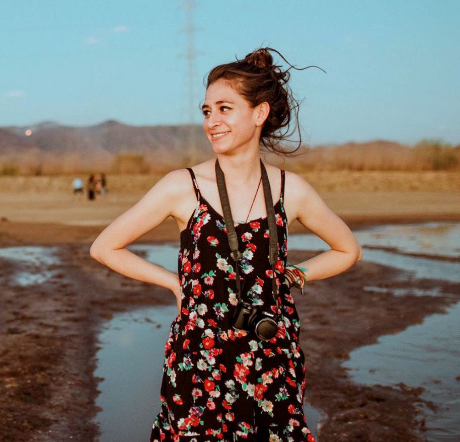 A girl with a flower sundress and a camera stands in a muddy field.