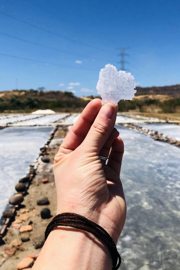A hand holding up a large crystal of salt in front of a salt evaporation pool.