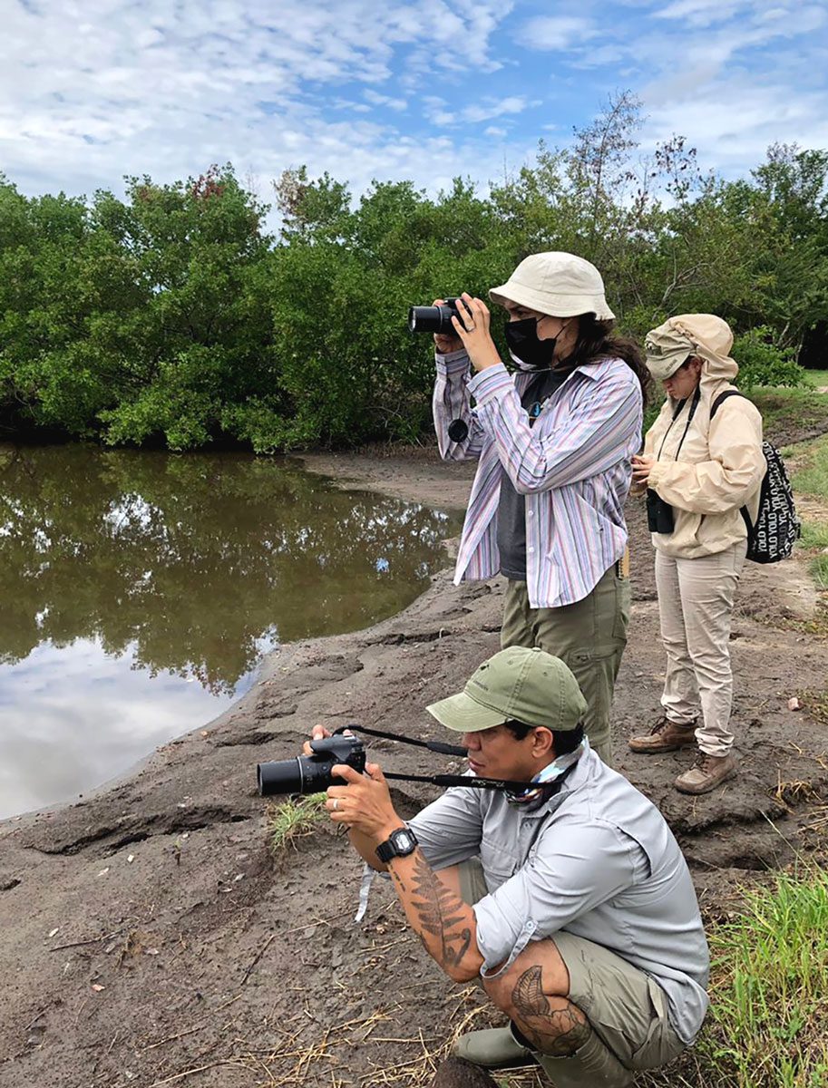 Three people standing by water with cameras and binoculars.