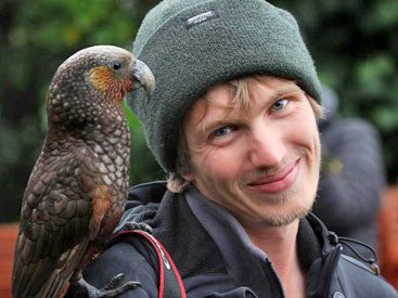 Smiling man with green hat and a large, dark-green parrot on shoulder.