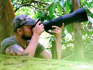 Man with camo baseball hat looking through a camera with a large lens, in the forest.