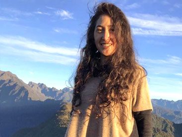 A long, brown-haired woman stands with mountains in the background.