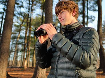 Young man with puffer jacket and binoculars, in the woods.