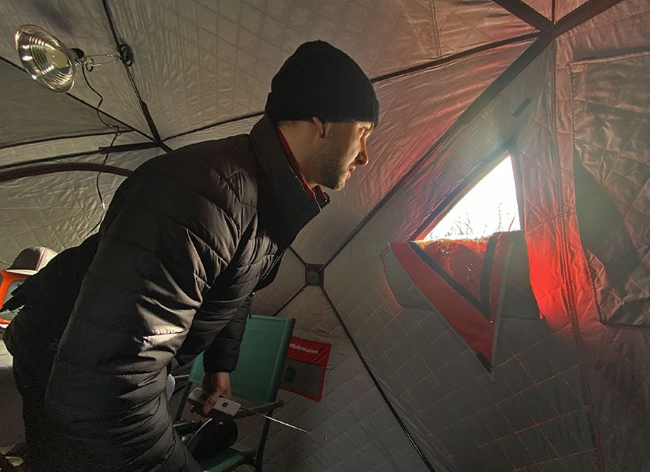 Man in a black coat and cap inside a tent, looking out a small window.