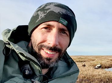 Bearded man with beanie smiles at camera, tundra in background.