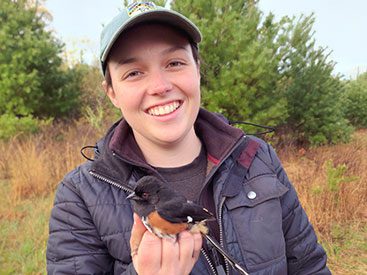 Young woman in baseball cap and jacket, smiles at the camera and holds a bird.