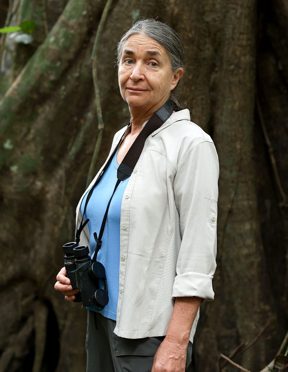 Una mujer mayor con una camisa blanca y binoculares alrededor del cuello está de pie junto a las raíces de un árbol gigante.