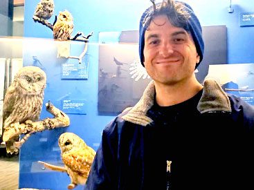 Smiling man in blue beanie and owl exhibit behind him.