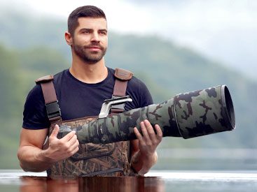 Dark-haired man with neat haircut and beard stands in waist-high water and holds a ridiculously large-lensed camera.