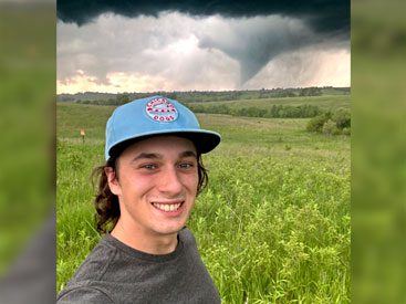 Young man in blue baseball cap smiles at camera, with tornado in far distance.