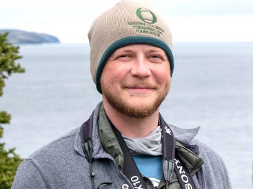 Man with beanie smiles at camera with body of water in background.