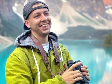 Man with a backwards baseball cam, chartreuse jacket and binoculars, stands with a mountain lake background.