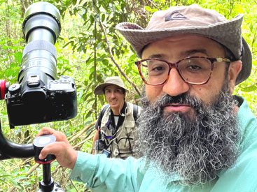 Bearded man with glasses and wide-brimmed cap, stands in the woods with a large-lensed camera, and another man in the background.