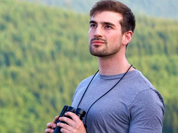 Man with gray T-shirt and binoculars stands with mountainous forest in background.