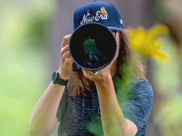 Man with baseball cap and longish hair looks through the camera lens, faces the photographer and is almost hidden behind large camera lens